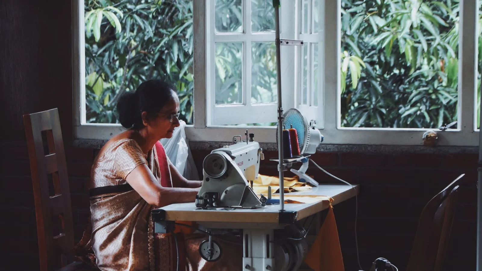 Indian woman in a sari stitching a garment on a sewing machine beside a window in a small workshop
