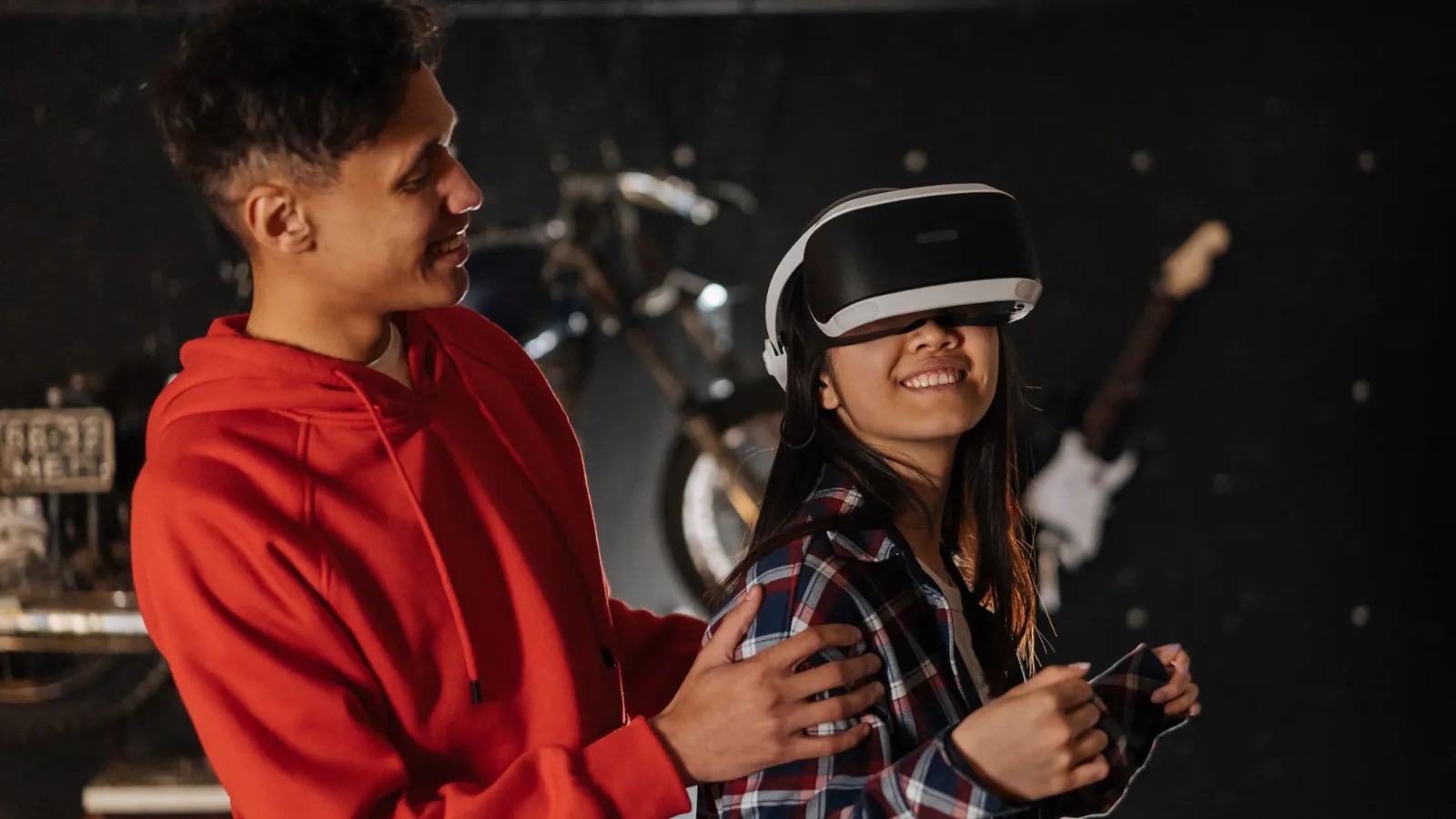 Two young friends at a gaming session, one wearing a virtual reality headset while the other helps him