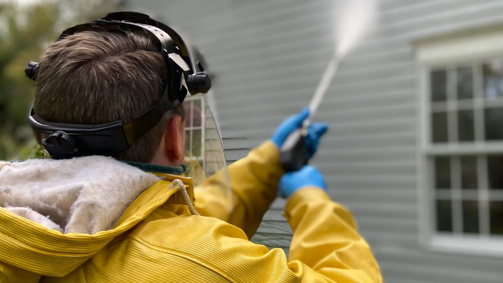 Back view of a worker in a yellow rain jacket and protective gear pressure washing the siding of a house